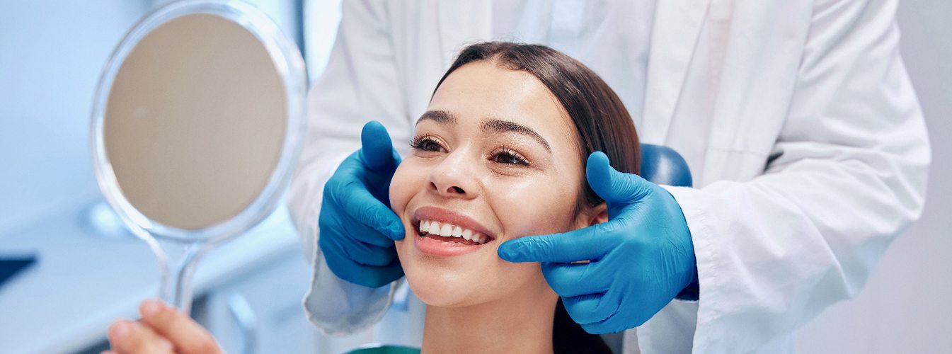 a woman smiling in a handheld mirror while sitting in a dental chair